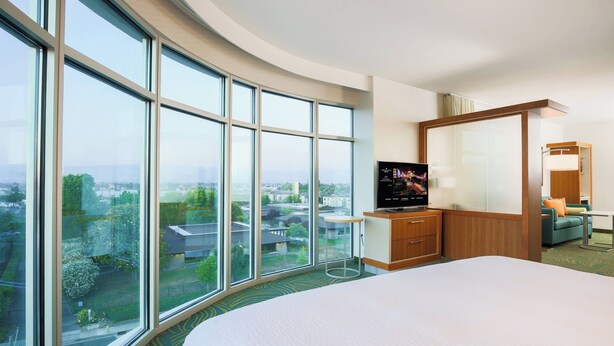 Habitación de hotel con una cama grande, un escritorio, un televisor y vistas amplias de la ciudad a través de grandes ventanales.
