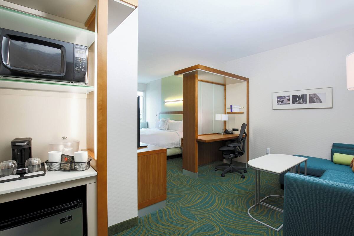 A hotel room with a kitchenette, a bed, a desk, a chair, a small table, and a sofa.