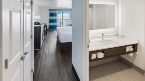 A hotel room with a large bed, a bathroom with a double sink, and a view of the sky through the balcony curtains. - Tropical Seas Hotel (Myrtle Beach)