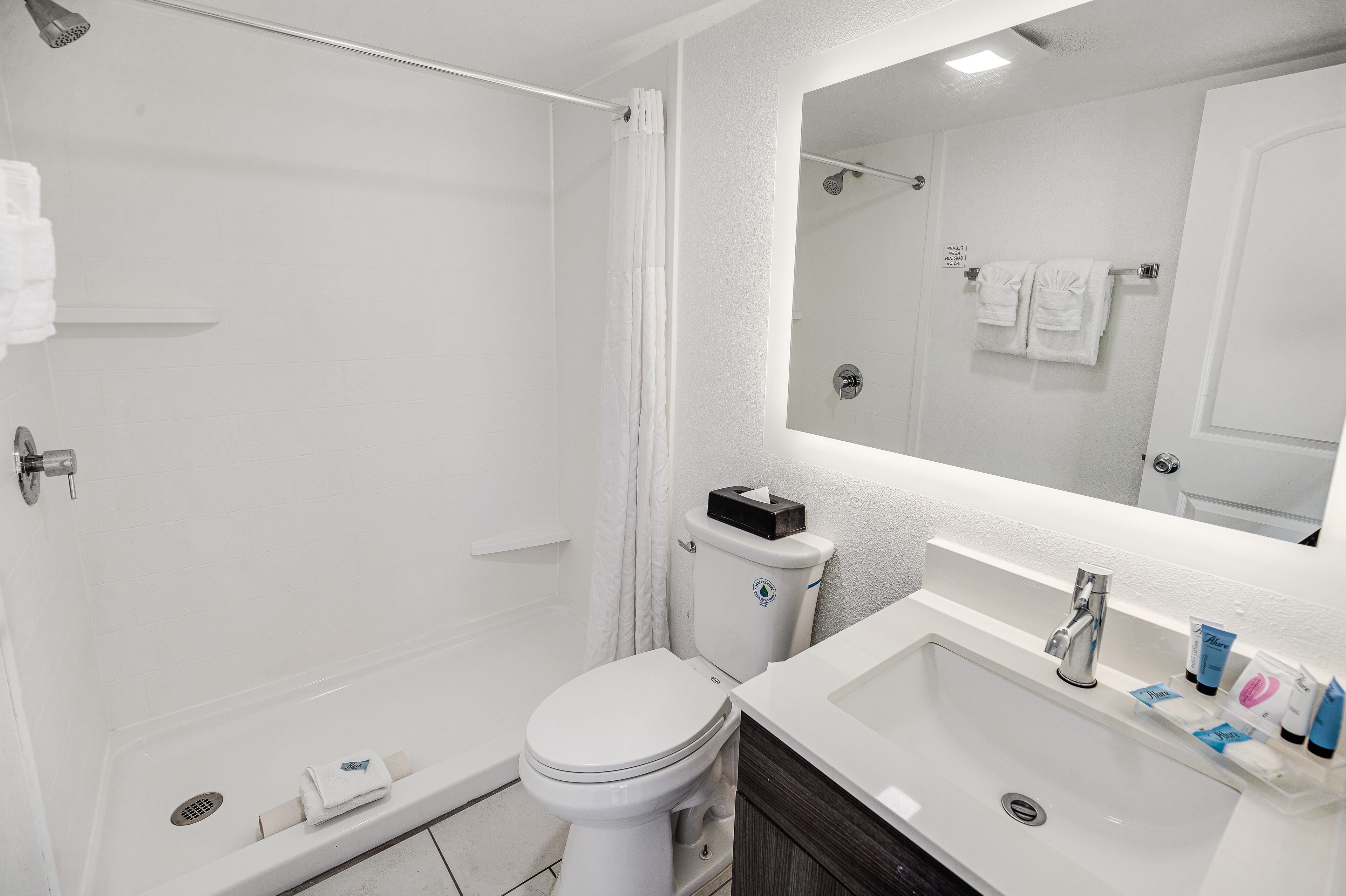 A bathroom with a white toilet, a shower, a sink with toiletries, and a mirror.