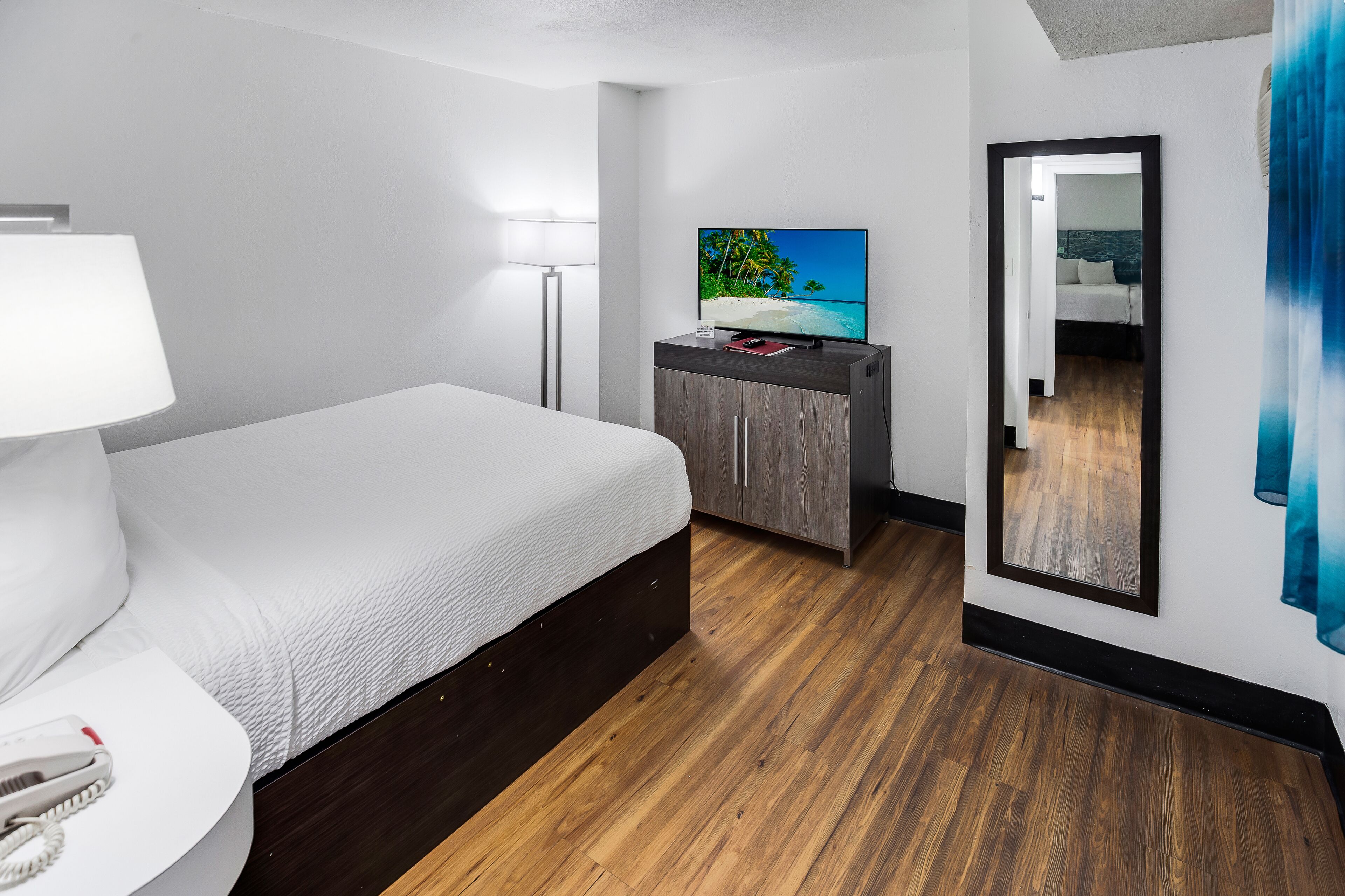 A modern hotel room with a wooden floor, a bed with white bedding, a flat-screen TV on a wooden cabinet, a bedside lamp, and a mirror on the wall.