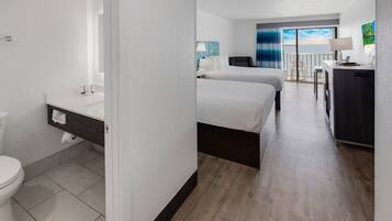 A hotel room with a bathroom, two beds, a desk, and a view of the ocean.