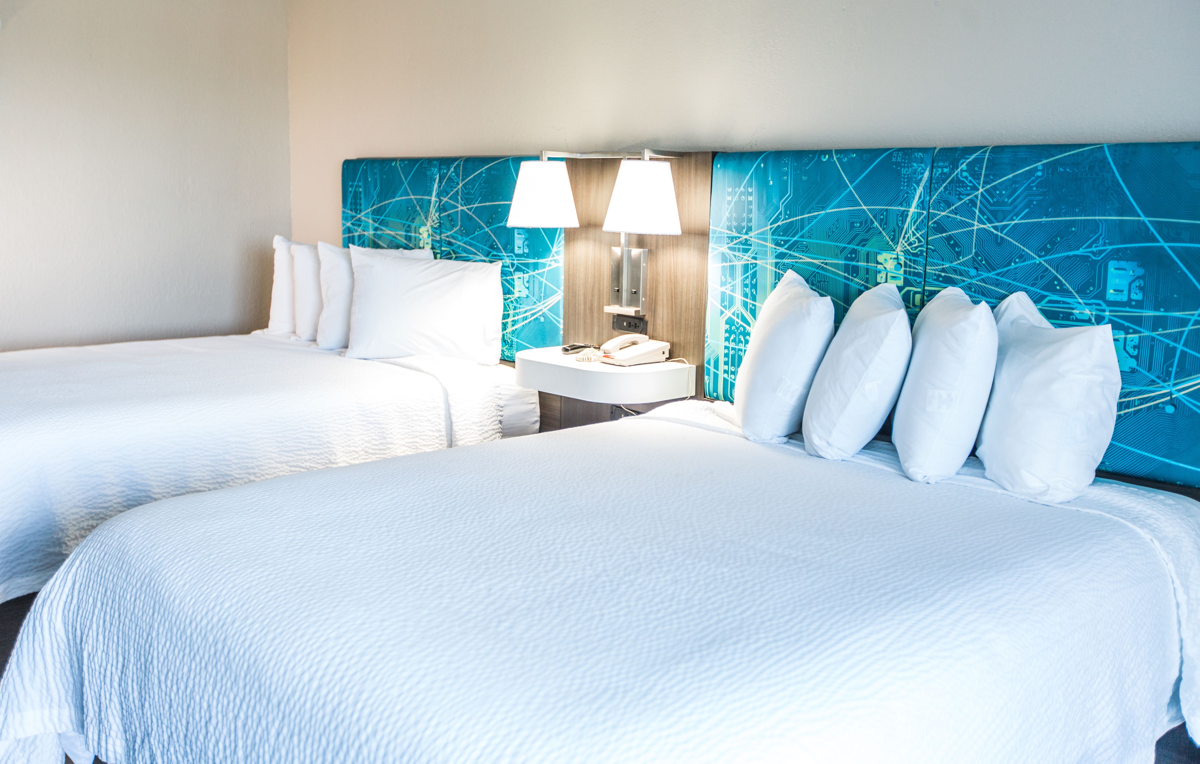 A hotel room with two beds, a nightstand with a lamp, and a wall with a blue circuit board design.