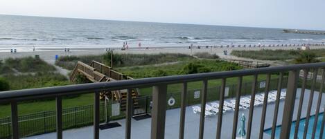 A balcony with a pool area, overlooking a beach with people and a pier.
