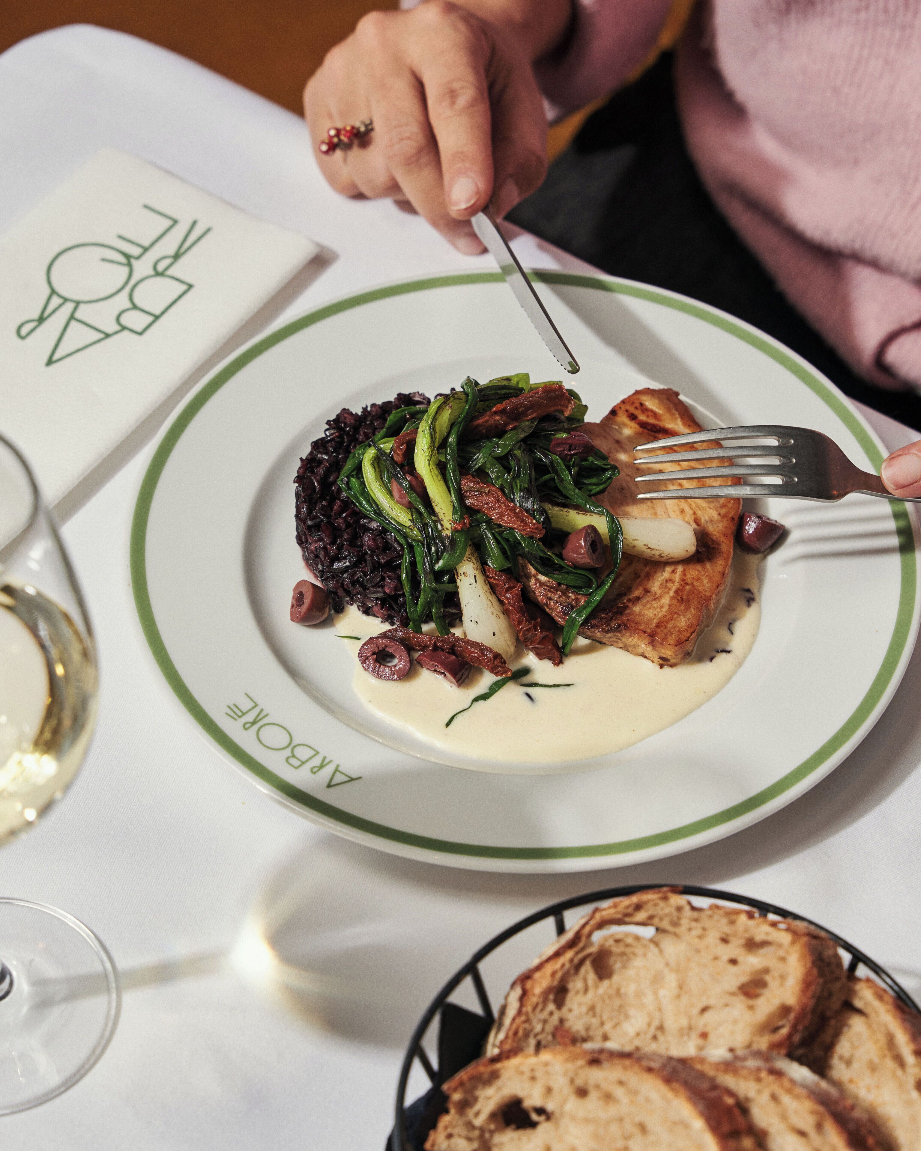 Une assiette de nourriture, accompagnée d’une fourchette et d’un couteau, un verre de vin blanc et un panier de pain, le tout posé sur une table.