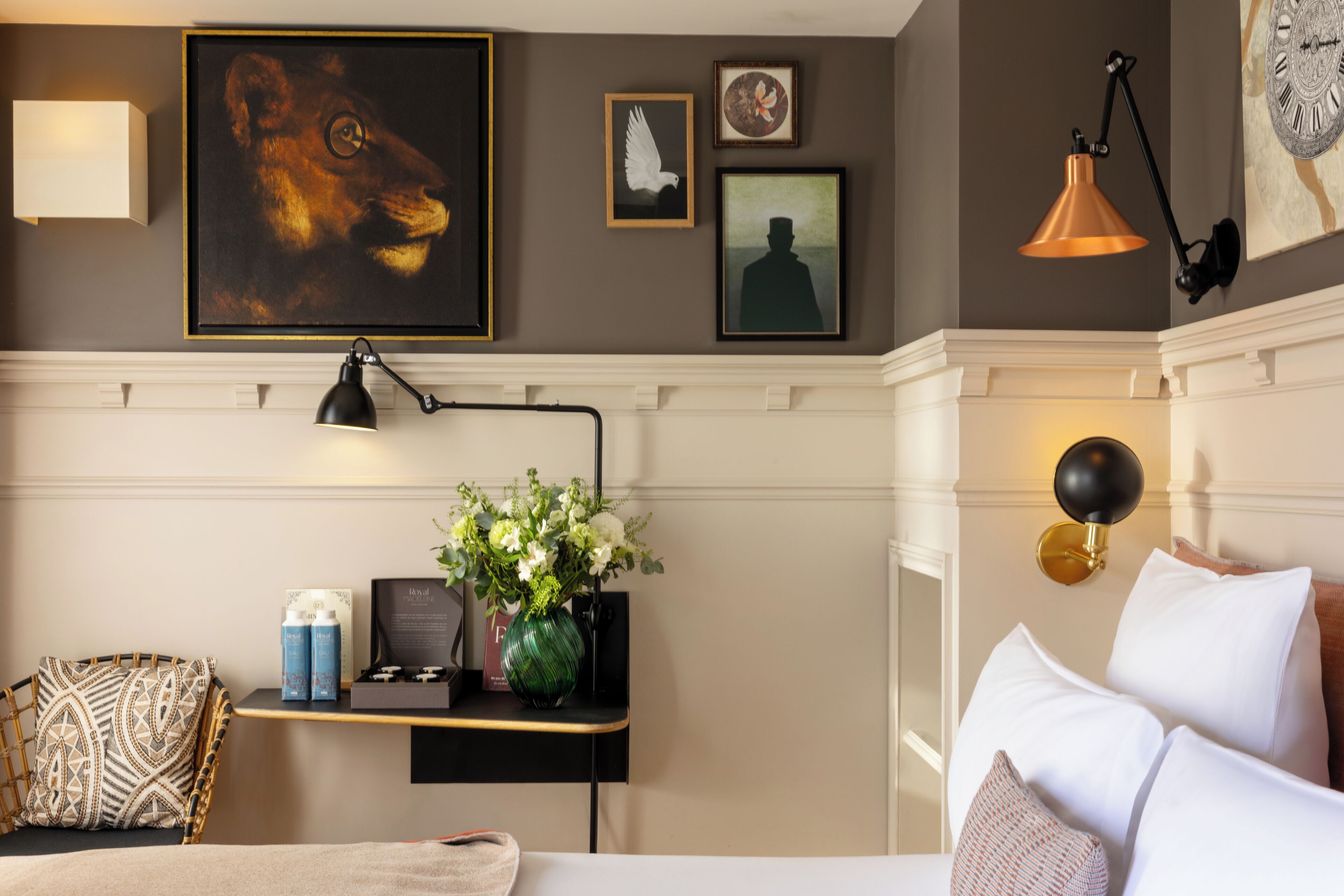 A hotel room with a bed, a bedside table with a lamp, a chair, a vase with flowers, and wall art.