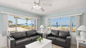 Quarto com dois sofás de couro preto, uma mesa de centro branca, um ventilador de teto e grandes janelas com vista para uma praia.