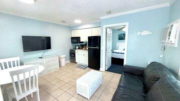 Un espacio habitable compacto con kitchenette, una zona de comedor con mesa y silla blancos, un sofá de cuero negro y un televisor montado en la pared.