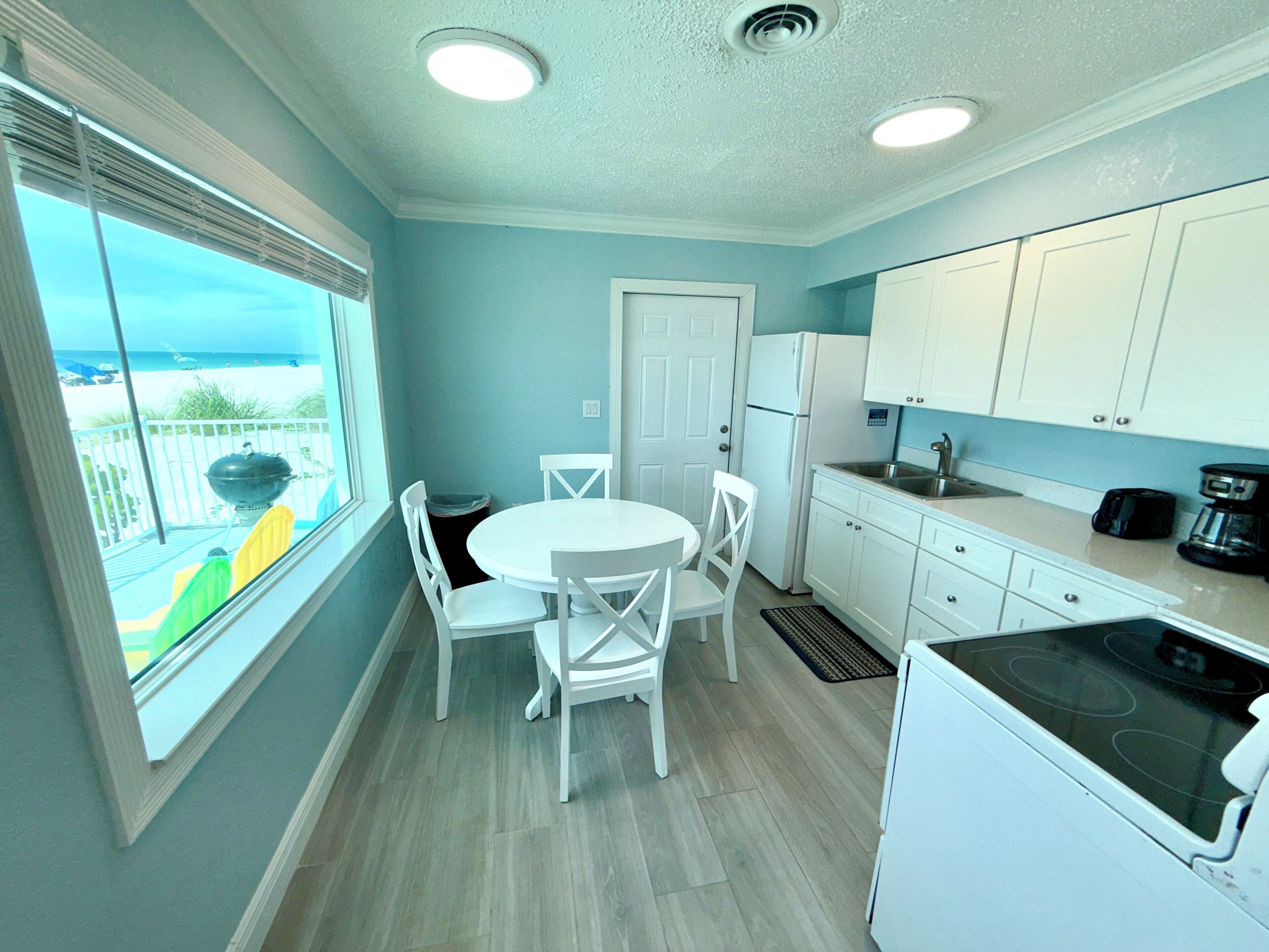 A compact kitchen with white cabinets, a white refrigerator, and a stove. There is a window with a view of the beach.