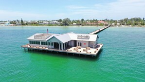 Uma casa flutuante à beira-mar com um deque coberto, área de estar externa e um mastro.