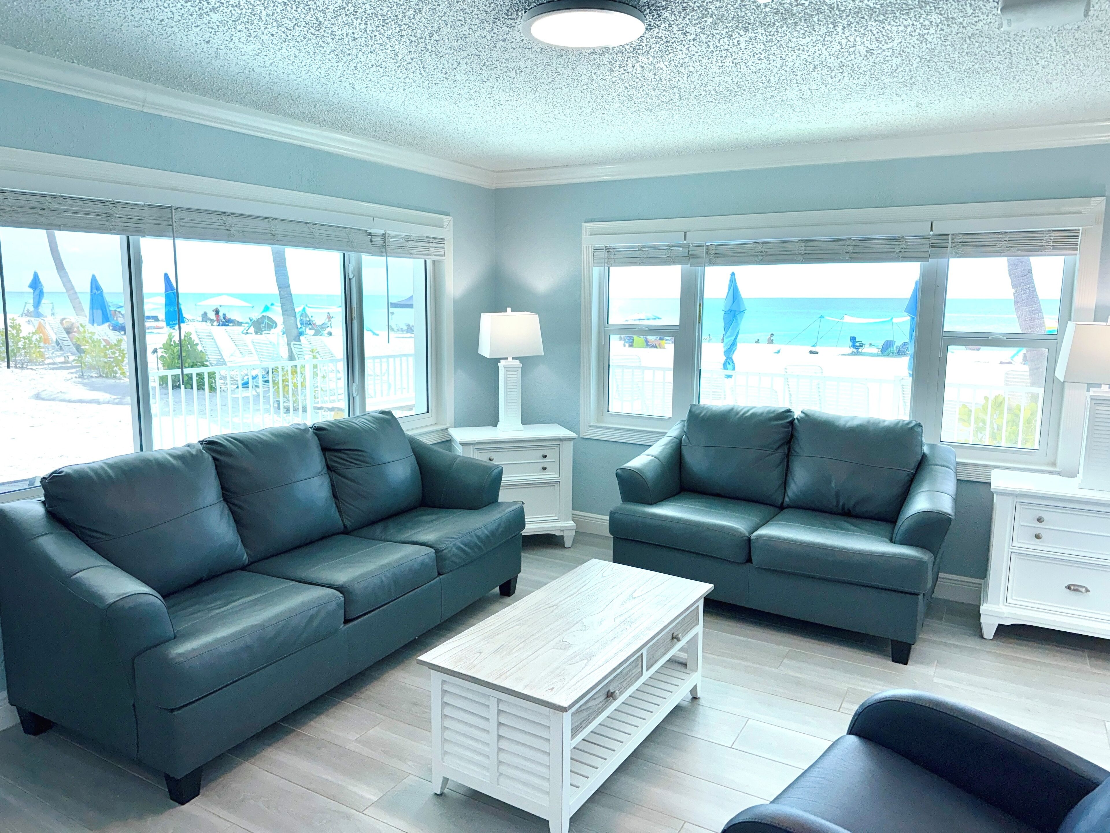 A room with two dark green leather sofas, a white coffee table, and a view of the beach through large windows.