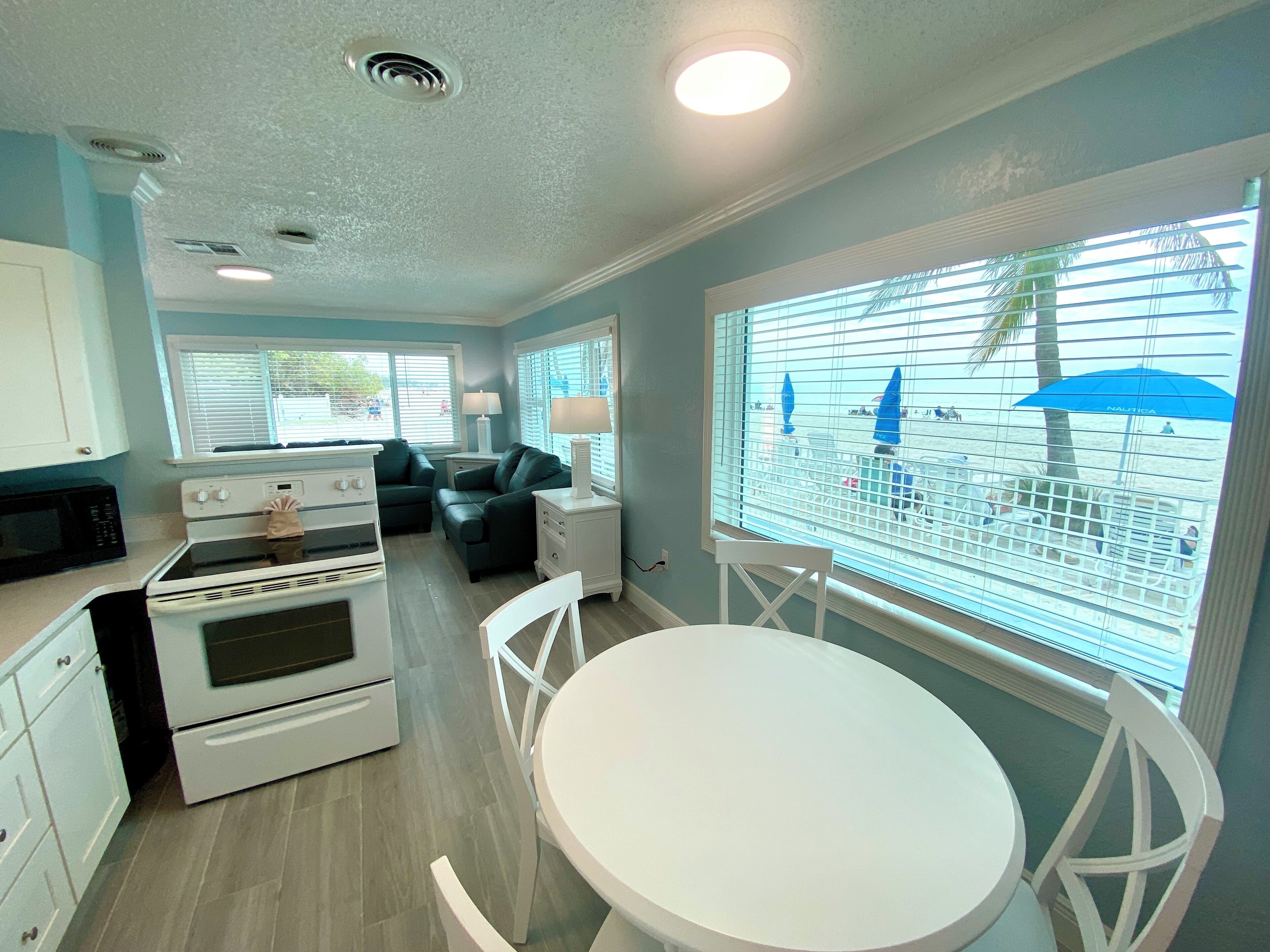 A kitchen with white appliances, a dining area with a white table and chairs, and a view of a beach through the window.
