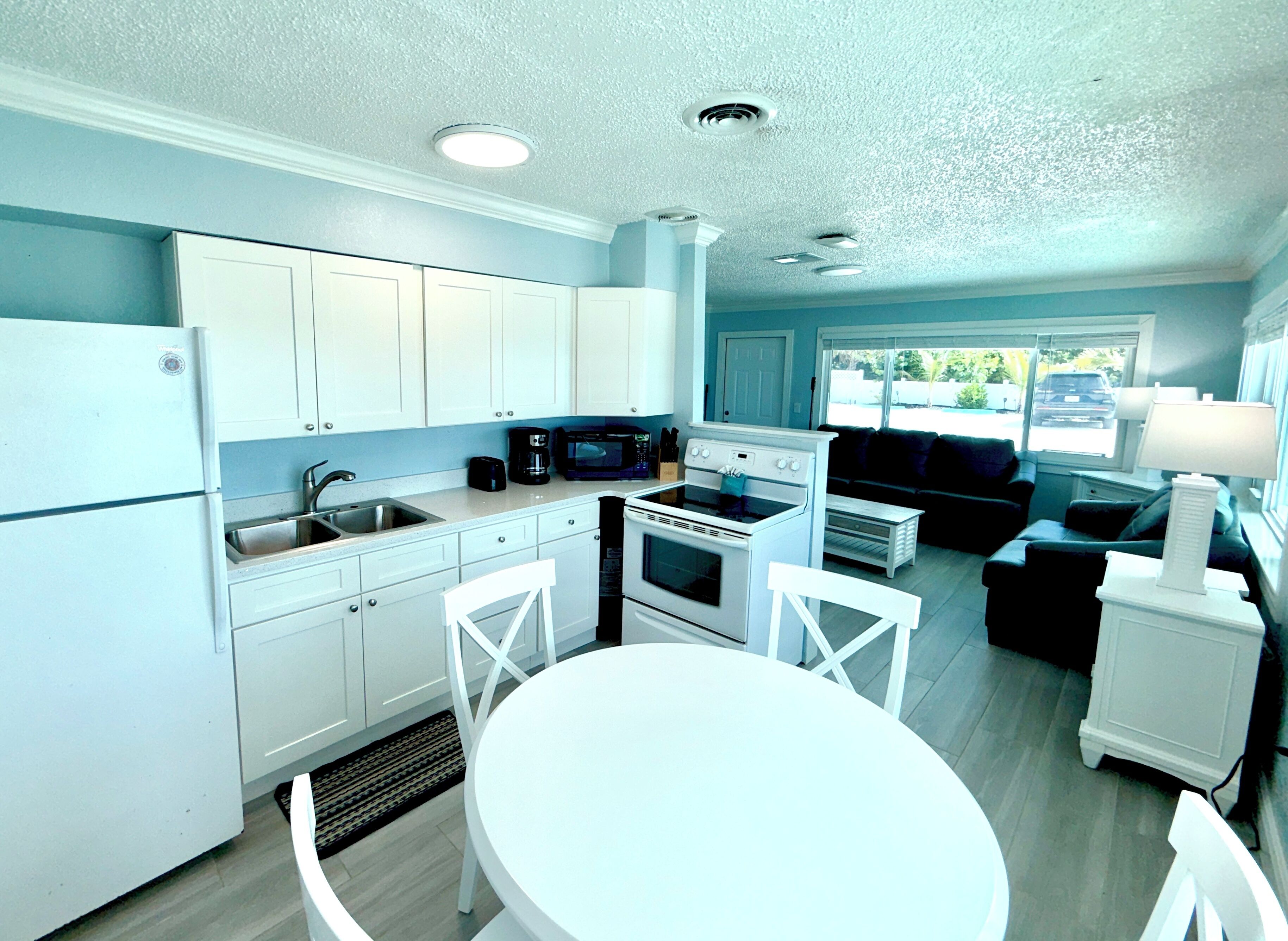 A modern kitchen with white cabinets, a refrigerator, a sink, a microwave, and an oven. There is a dining table with chairs and a sofa in the living area.