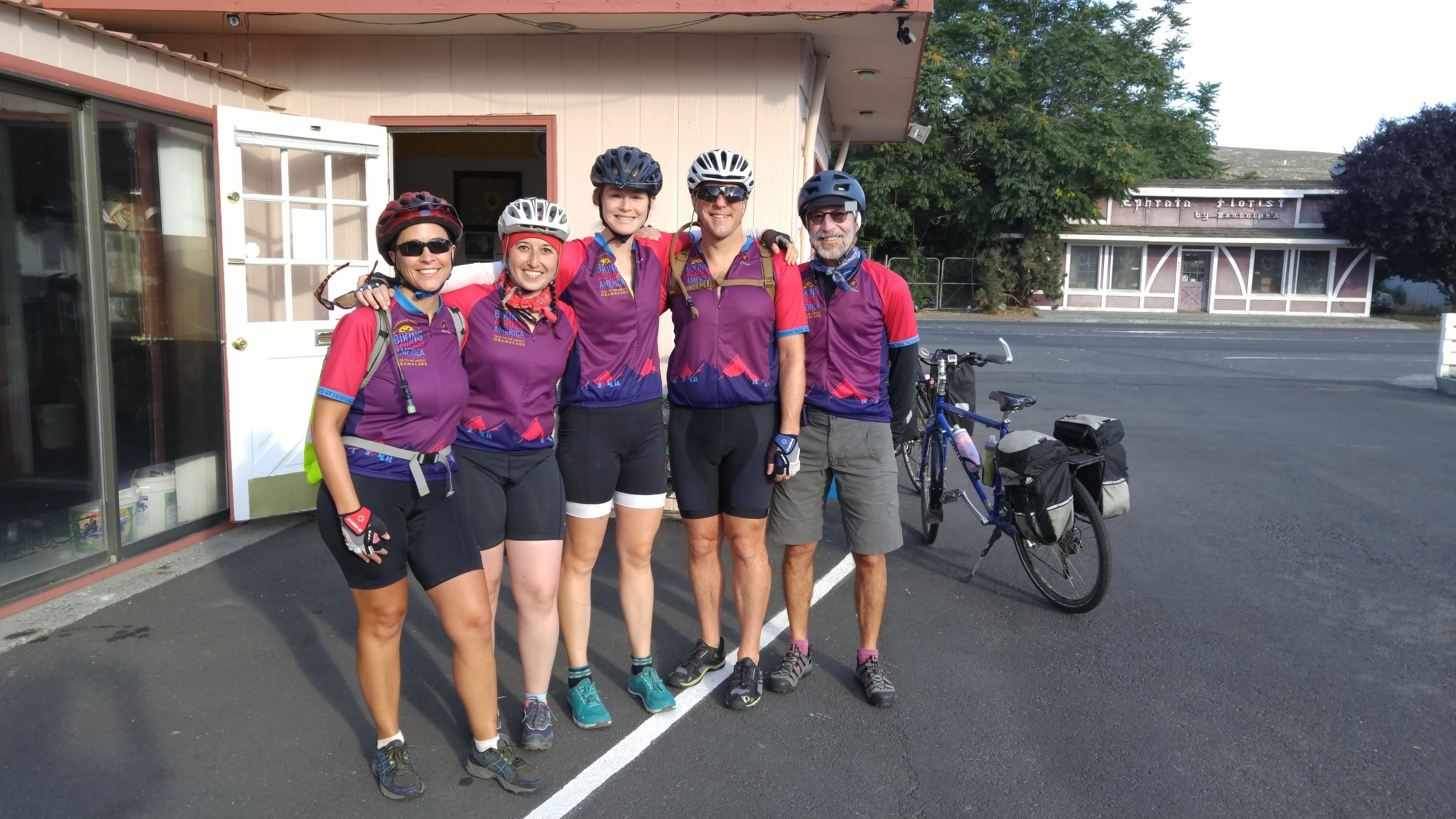 Un grupo de personas vestidas para andar en bicicleta, de pie frente a un edificio con una bicicleta estacionada cerca.