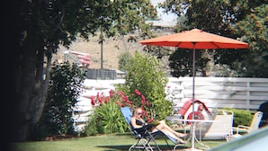 A person relaxing on a blue lawn chair under an orange umbrella, with a white fence and greenery in the background. - Ephrata Inn Motel (Ephrata)