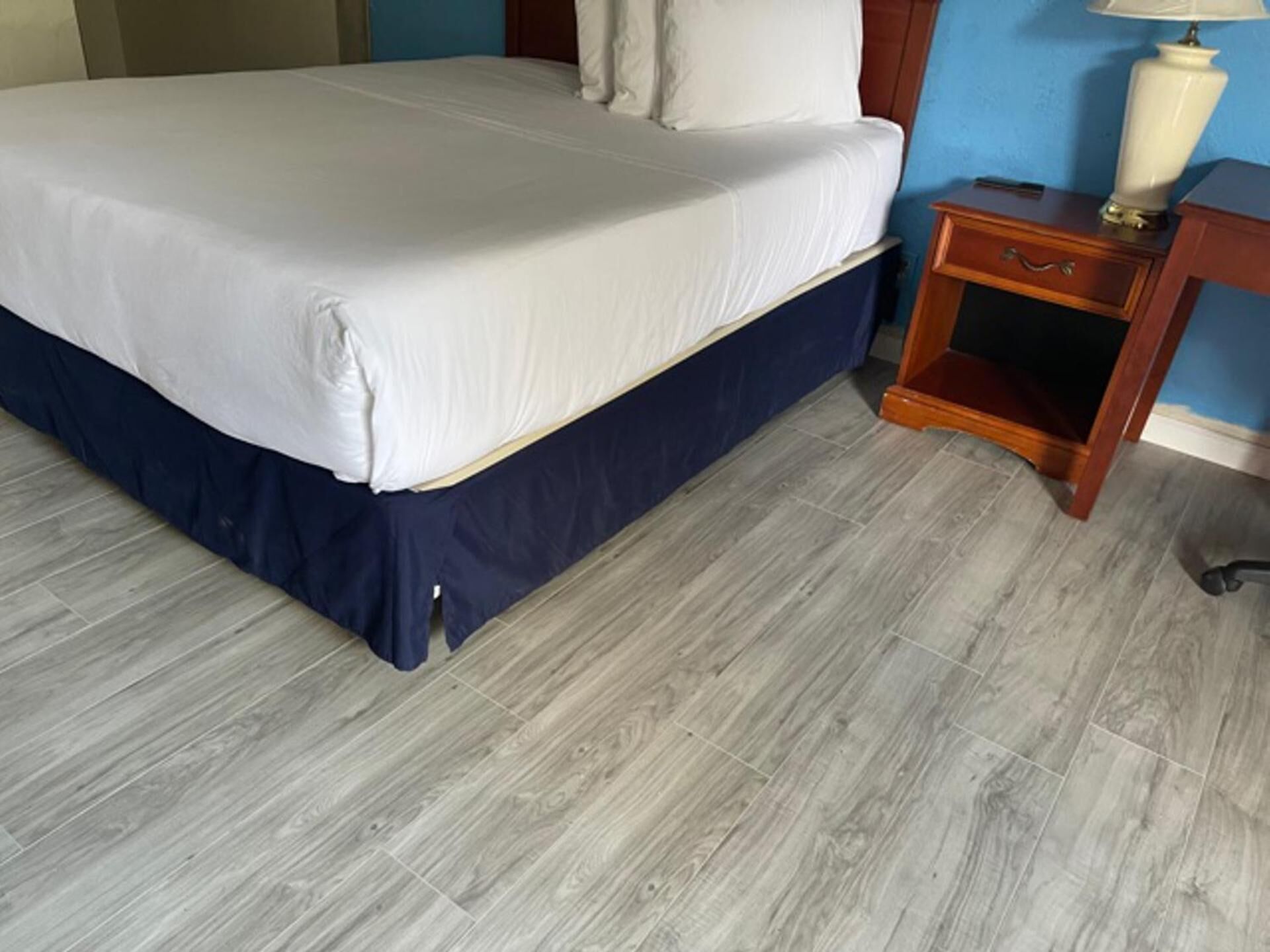 A bed with a white mattress and navy blue skirt, a wooden nightstand with a drawer, and a lamp.
