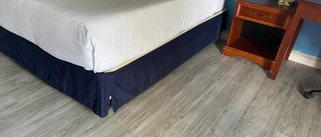 A bed with a white mattress and navy blue skirt, a wooden nightstand with a drawer, and a lamp.