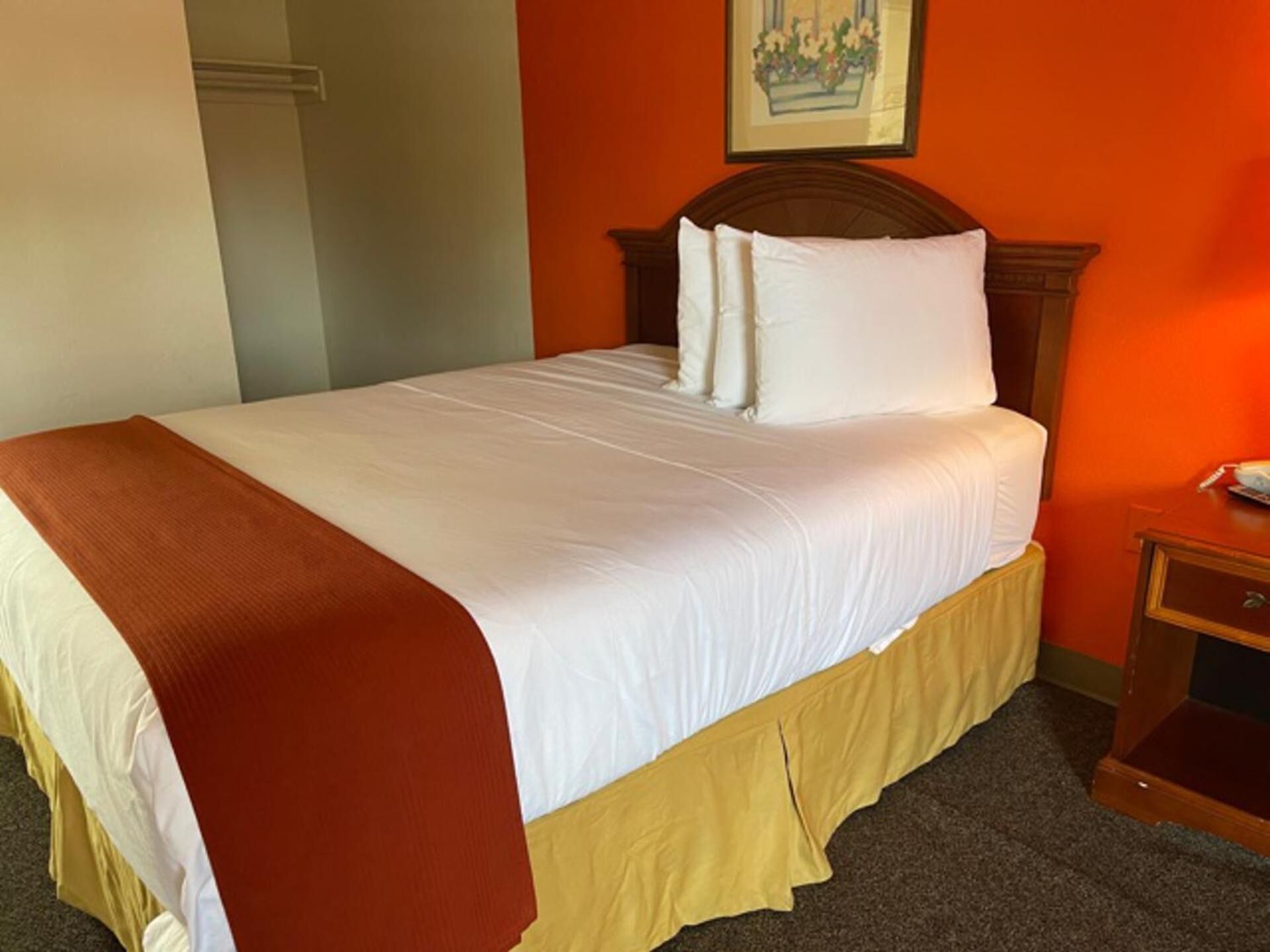 Una cama bien hecha con sábanas blancas y un plaid marrón, un cabecero de madera y una mesita de noche con una lámpara.