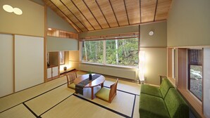 Ett traditionellt japanskt rum med tatamimattor på golvet, ett lågt bord med kuddar, en grön soffa och ett stort fönster med utsikt över träd.