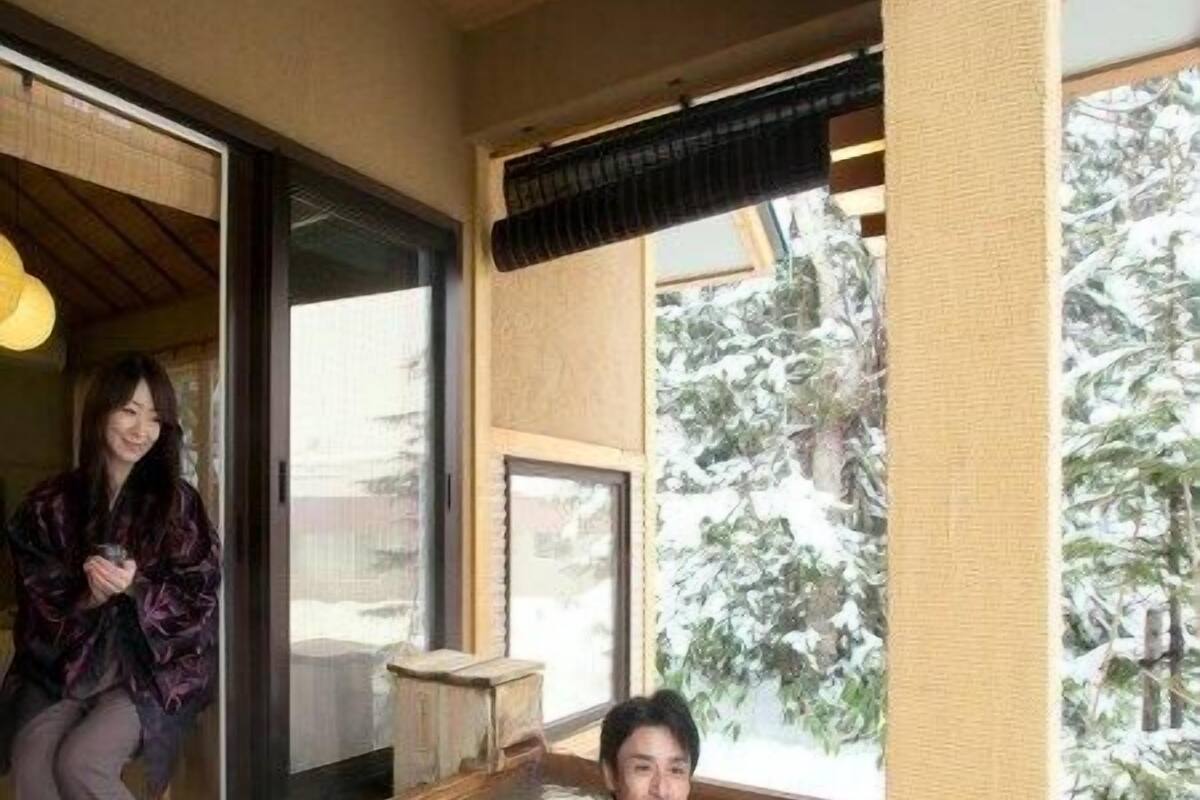 A traditional Japanese onsen with a wooden bath, a window with a view of snow-covered trees, and a person sitting outside the bath.