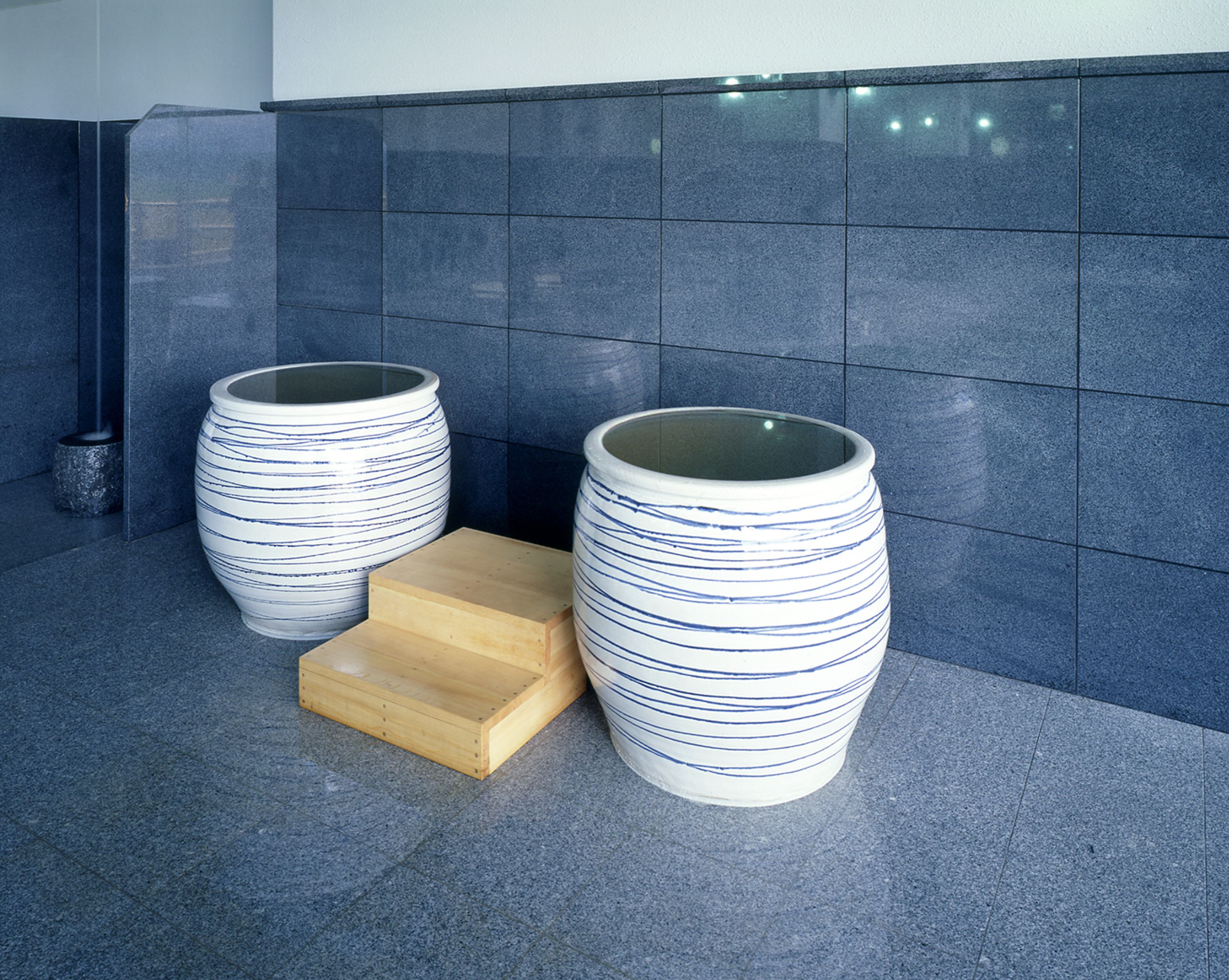 Two large white vases with blue stripes on a tiled floor, a wooden stool, and a dark blue tiled wall.