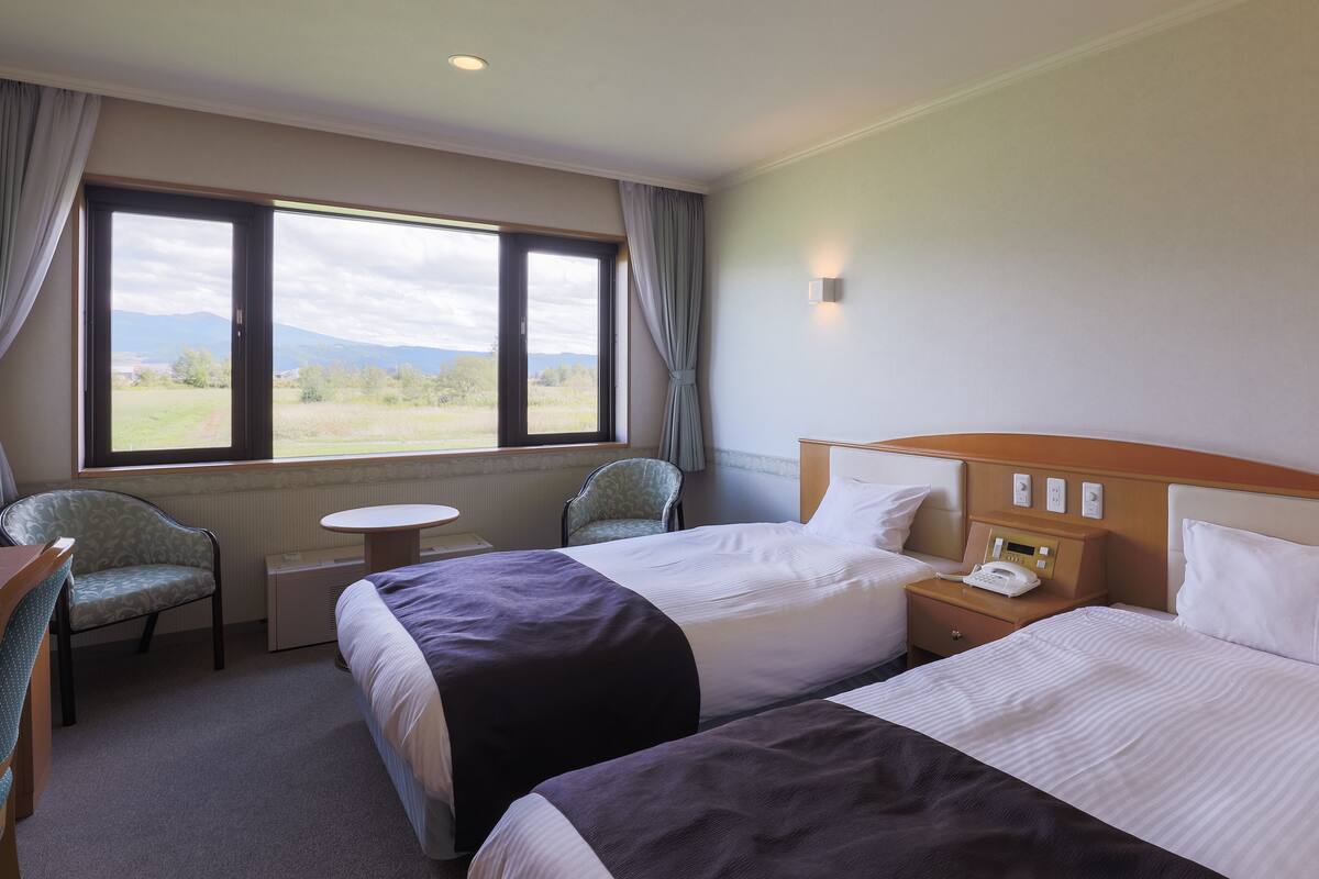 Une chambre d’hôtel avec deux lits, une petite table, une chaise, une fenêtre donnant sur la campagne et une table de chevet avec un téléphone.