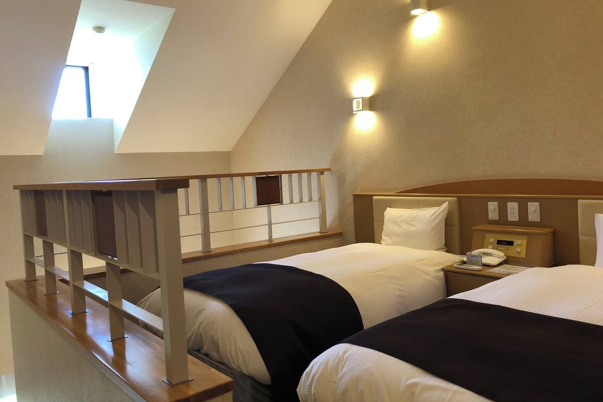 Une chambre d’hôtel avec deux lits, un escalier en bois, une lucarne et une table de chevet avec un téléphone et une horloge numérique.