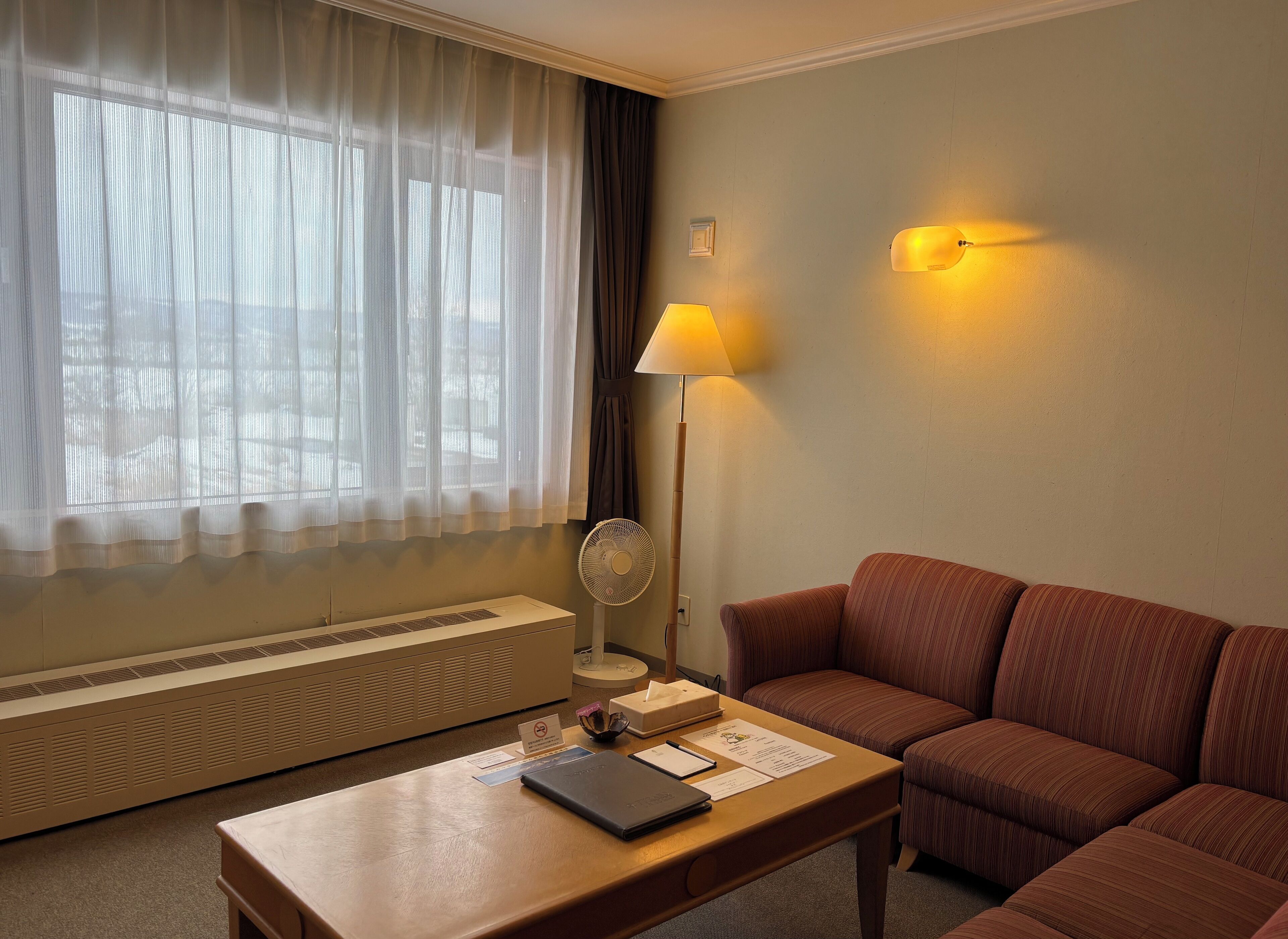 Quarto de hotel com sofá, mesa com um laptop, luminária, ventoinha e janela com cortinas leves.