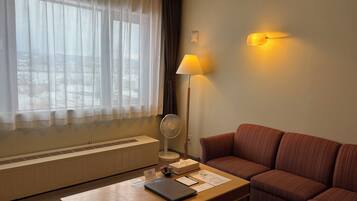 Quarto de hotel com sofá, mesa com um laptop, luminária, ventoinha e janela com cortinas leves.