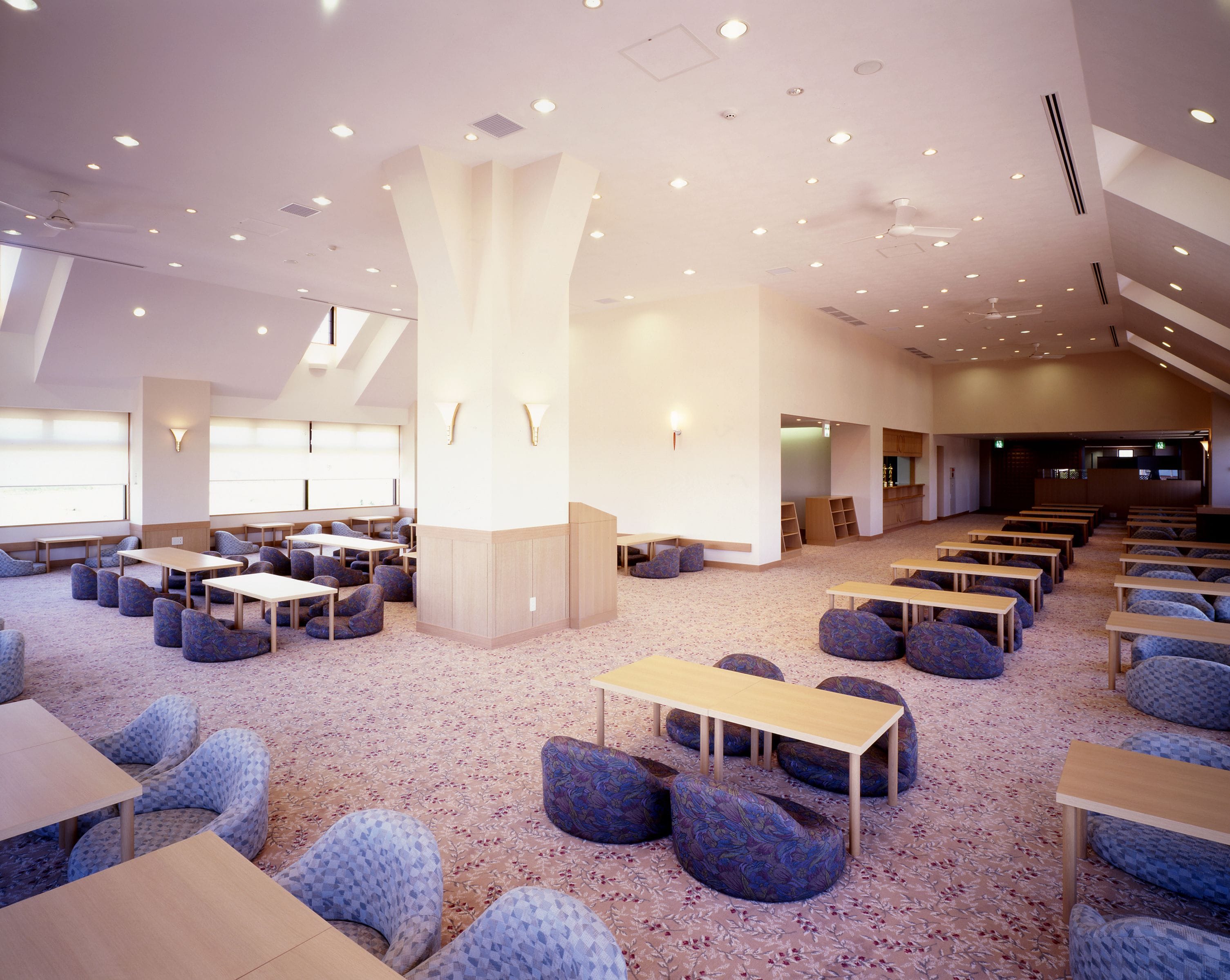 A spacious conference room with multiple tables and chairs, a high ceiling with recessed lighting, and a large pillar in the center.