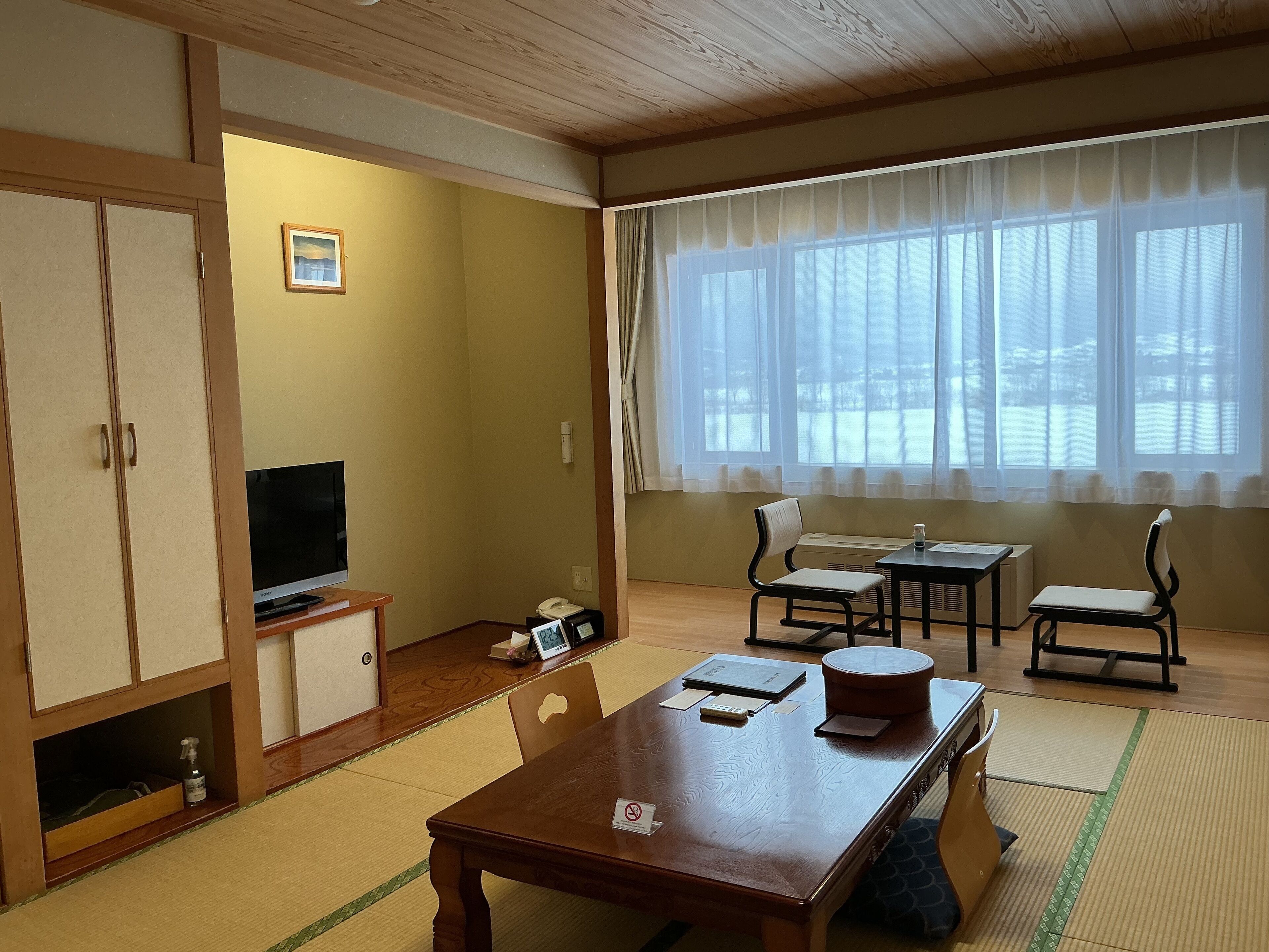 Una habitación con una mesa de madera, sillas, un televisor y una ventana con cortinas traslúcidas.