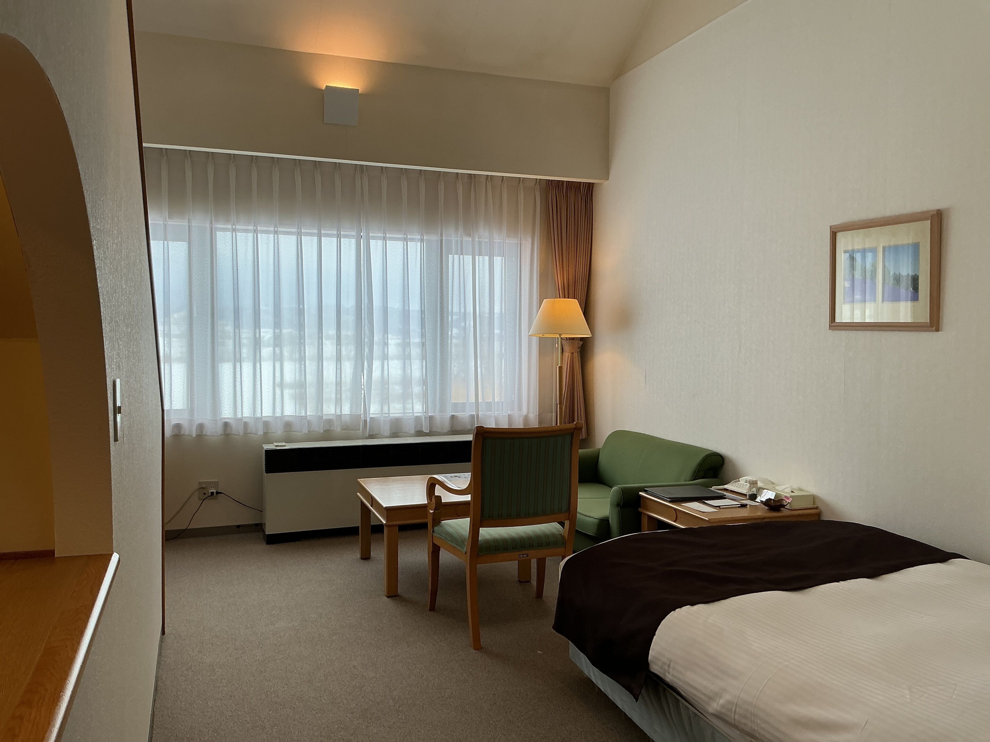 A hotel room with a bed, a desk with a chair, a lamp, and a window with curtains.