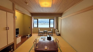 A traditional Japanese room with tatami flooring, a wooden table, chairs, a television, and a large window offering a city view.