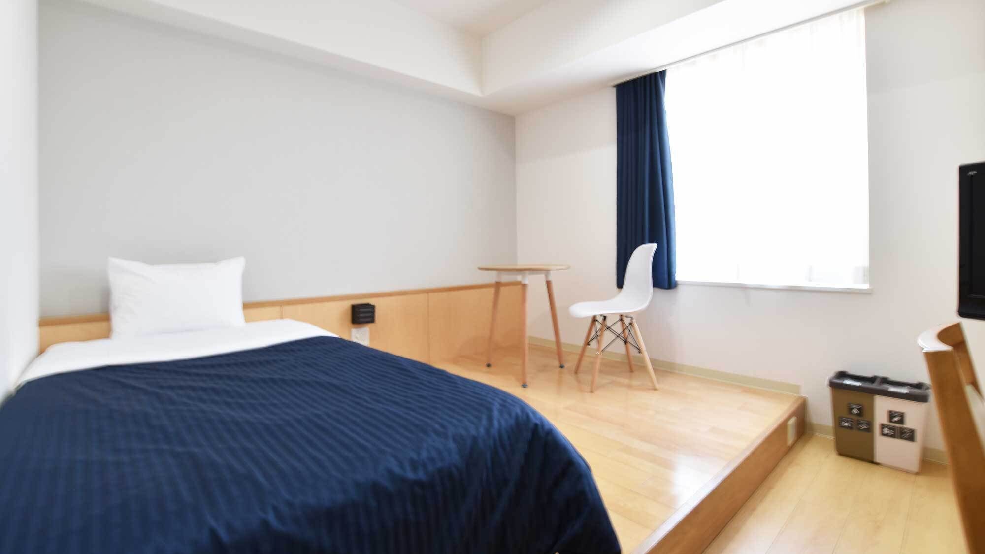 A hotel room with a bed, a small table, a chair, a window with curtains, and a trash bin.