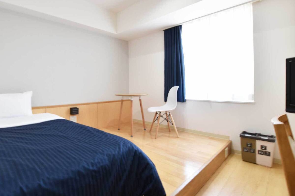 Una habitación de hotel con cama, una mesita, una silla, un ventanal con cortinas y un cesto de basura.