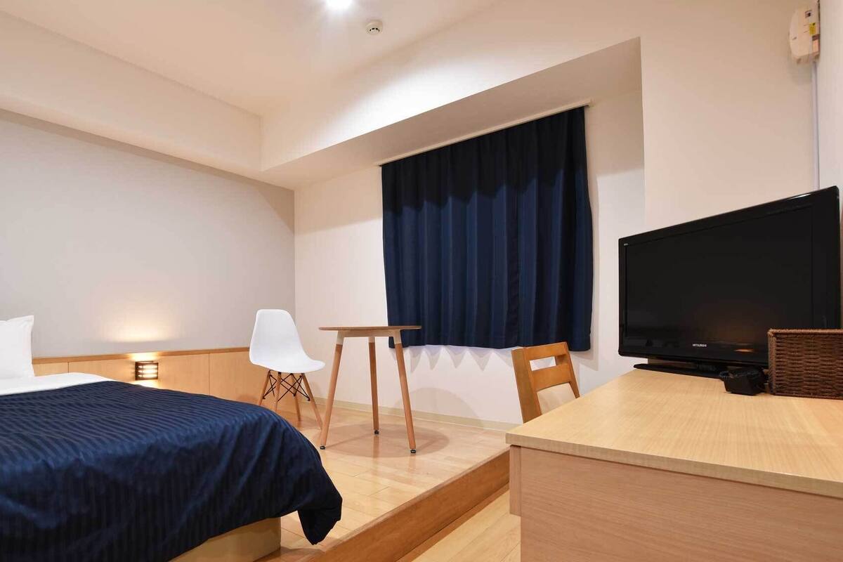 Una habitación de hotel con cama, escritorio, silla, televisor y ventana con cortinas.