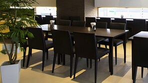 A conference room with multiple tables and chairs, a potted plant, and a view through the windows.