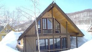 A wooden cabin with a gabled roof, large windows, and a visible attic space. - Niseko Hirafu Izumikyo Rental Cottage (Kutchan)