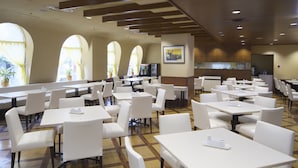 A spacious dining area with multiple tables and chairs, large windows with curtains, and a kitchen area in the background. - Hotel North City (Sapporo)