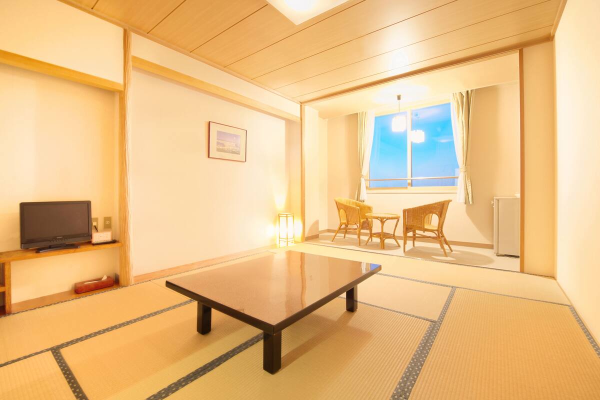 A traditional Japanese room with tatami flooring, a low table, a television, and a window with a view of the sky.