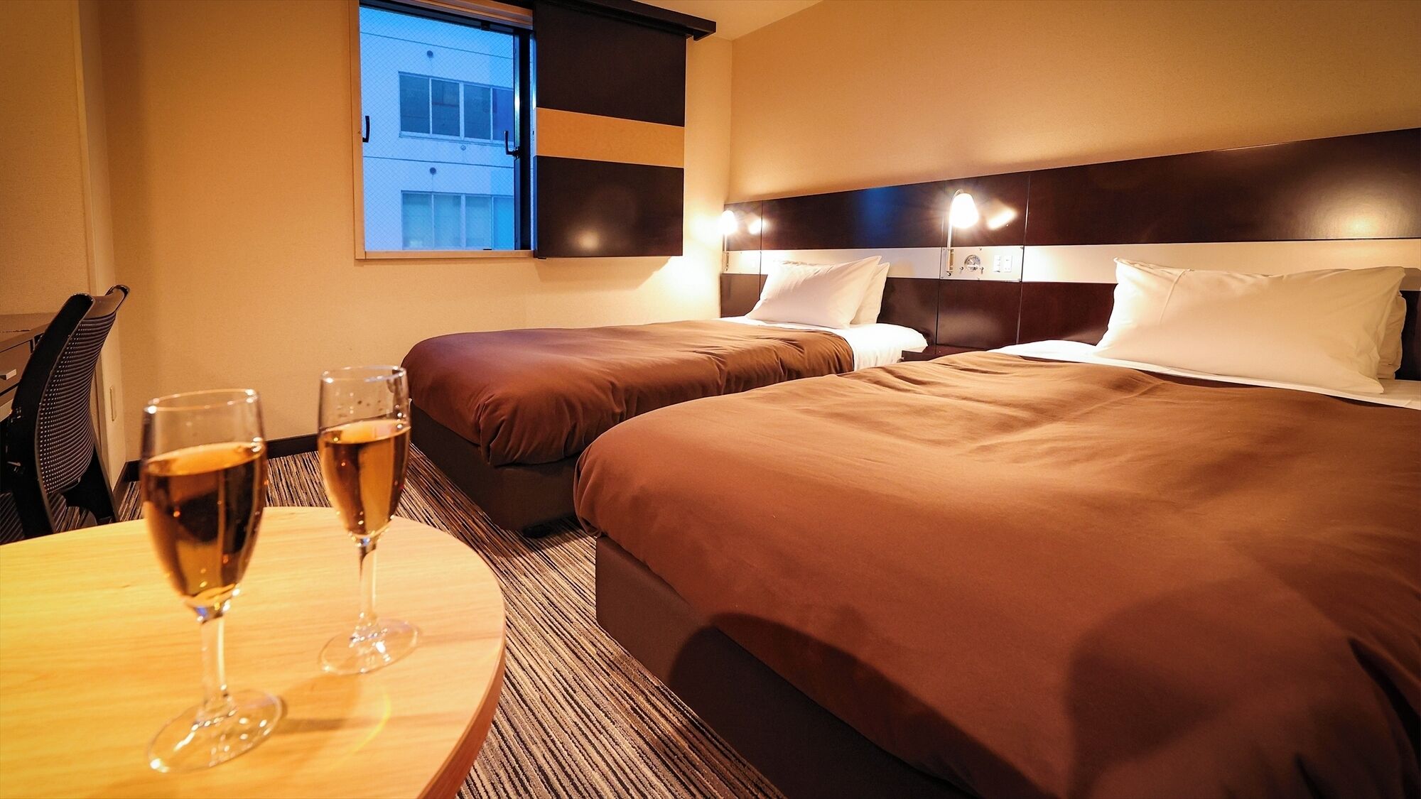 Ein Hotelzimmer mit zwei Betten, einem Tisch mit zwei Weingläsern und einem Fenster mit Blick auf ein Gebäude.