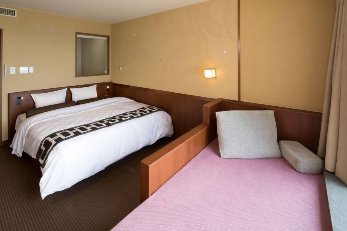 Habitación de hotel con una cama grande, una cama más pequeña, una mesita de noche, un televisor montado en la pared y un ventanal con cortinas.