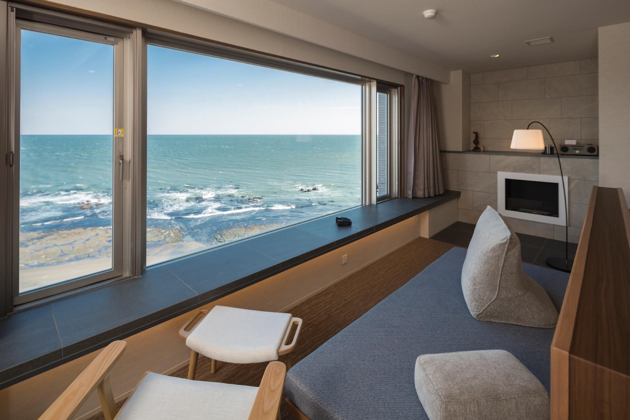A room with a large window overlooking the sea, a flat-screen TV, and a seating area with a bench and chairs.
