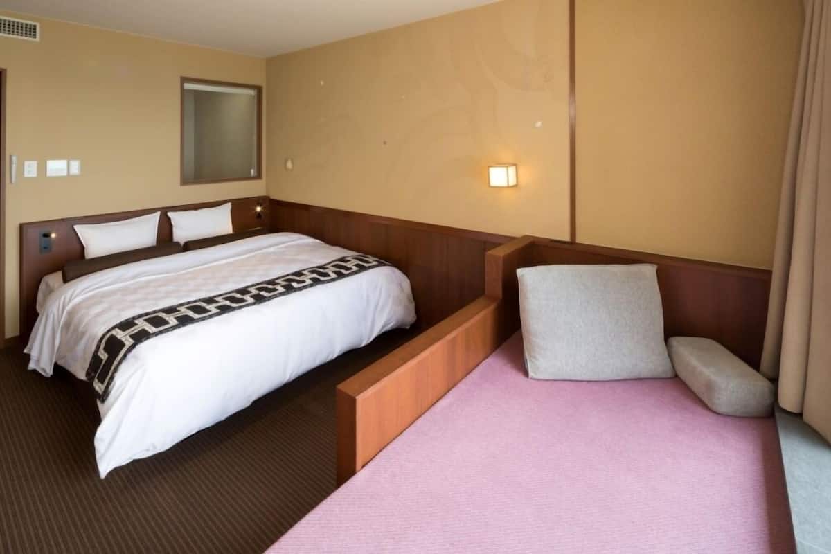 Una habitación de hotel con una cama grande, una cama más pequeña, una mesita de noche, un televisor montado en la pared y una ventana con cortinas.
