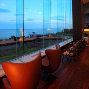 A room with a large glass window overlooking a body of water, wooden flooring, and modern chairs.
