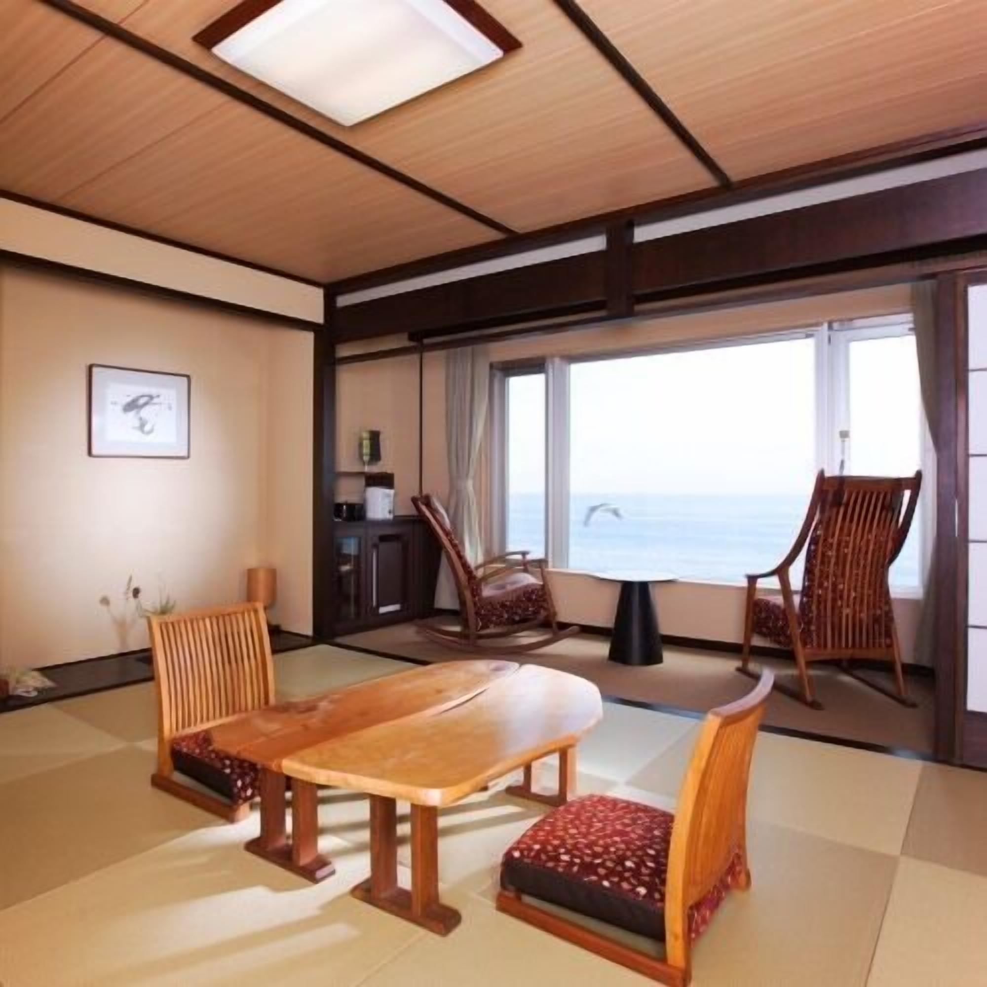 Una habitación con suelo de tatami, una mesa de madera, dos sillas, un mueble pequeño y vistas al mar a través de ventanas amplias.