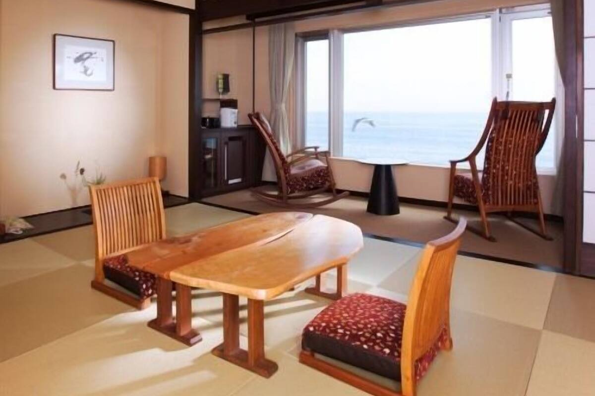Una habitación con suelo de tatami, una mesa de madera, dos sillas, un mueble pequeño y vistas al mar a través de ventanas amplias.