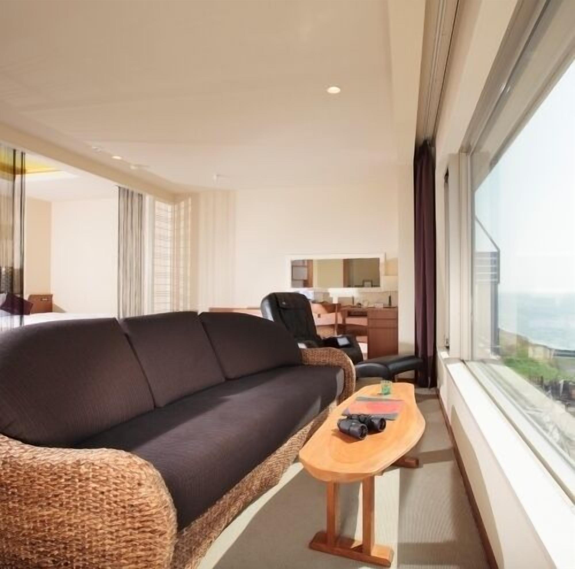 Habitación de hotel moderna con un sofá marrón oscuro, una mesa de centro de madera, un escritorio con una silla y un ventanal con vistas a la ciudad.