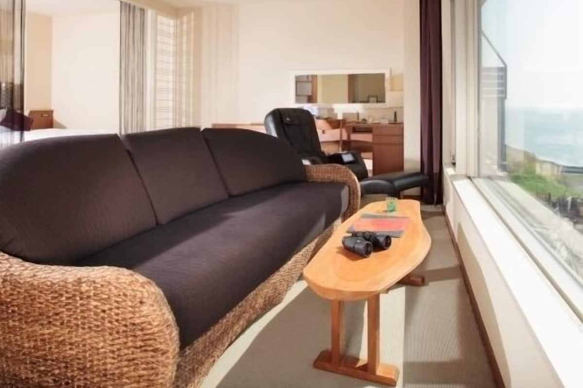 Habitación de hotel moderna con un sofá marrón oscuro, una mesa de centro de madera, un escritorio con una silla y un ventanal con vistas a la ciudad.