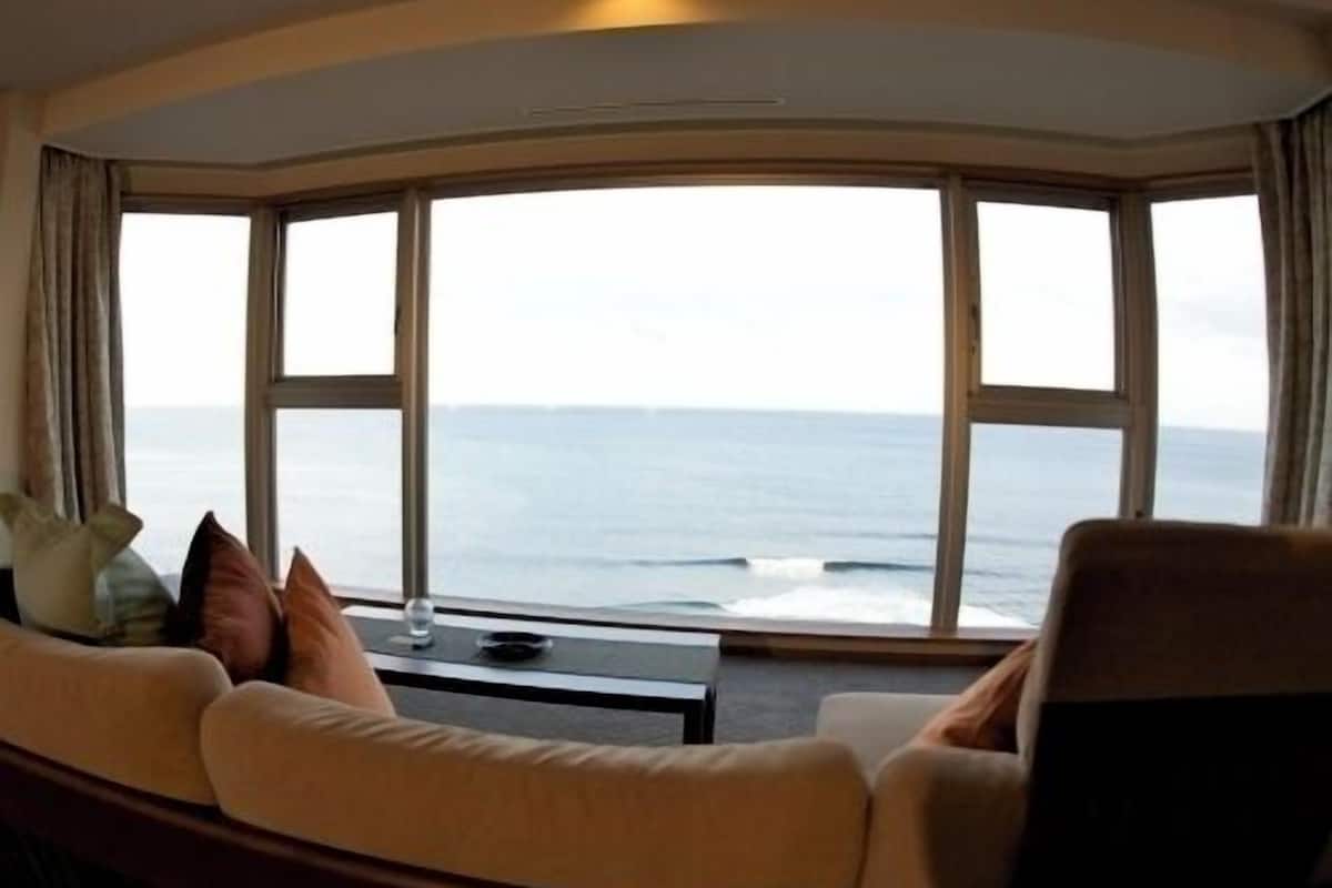 Una habitación con ventanal curvo con vista al mar, un sofá con cojines y una pequeña mesa.